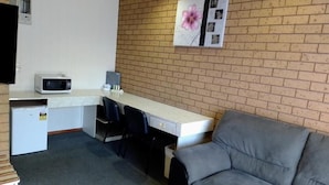 A compact room with a kitchenette, a grey sofa, and a brick wall with a floral artwork. - Country Home Motor Inn (Shepparton)