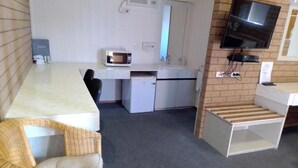 A compact hotel room with a kitchenette, a television, a microwave, a small refrigerator, a wicker chair, and a wooden bench. - Country Home Motor Inn (Shepparton)