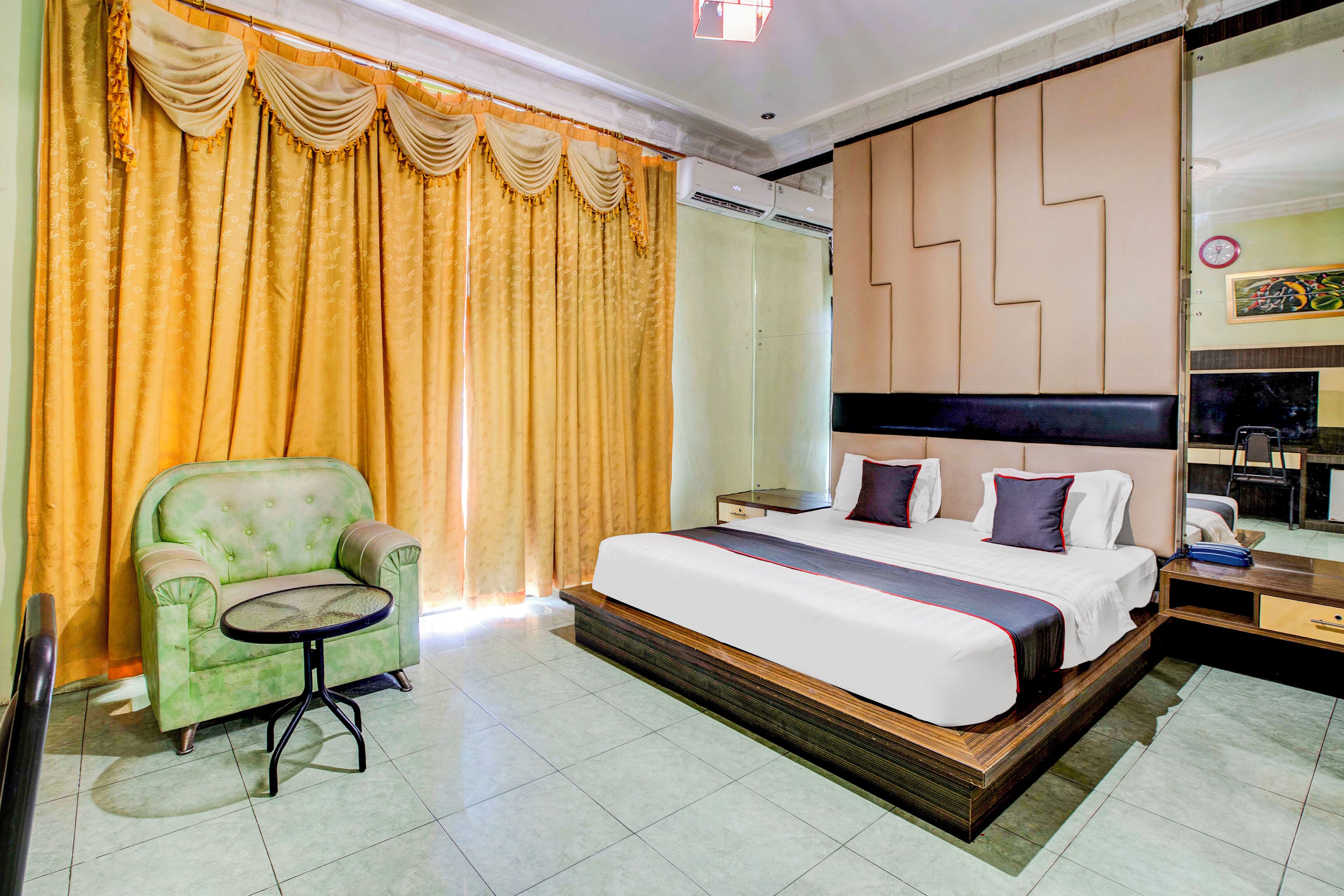 A hotel room with a large bed, a green armchair, a small round table, and a large mirror.
