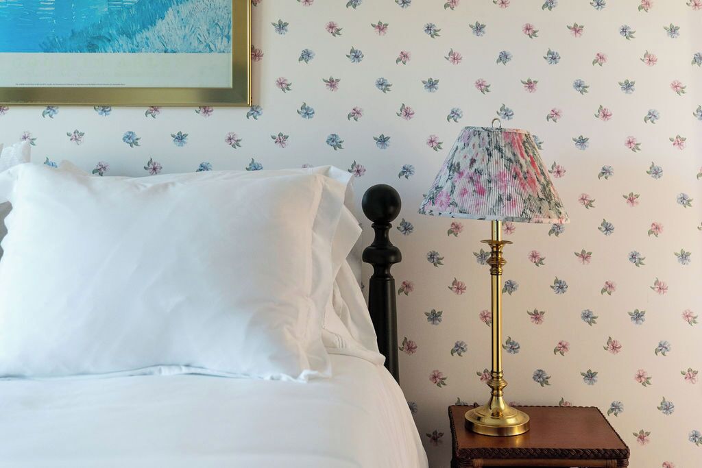 A neatly made bed with white linens, a floral-patterned lamp on a wooden nightstand, and a wall with a floral wallpaper pattern.