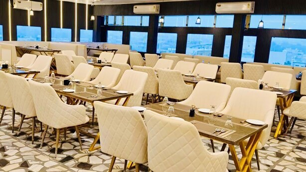 A conference room with multiple tables and chairs, large windows with a city view, and a patterned floor. - Hotel jageshwari inn (Gwalior)
