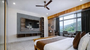A modern hotel room with a large bed, a flat-screen TV mounted on the wall, a ceiling fan, and a view of the outdoors through large windows.