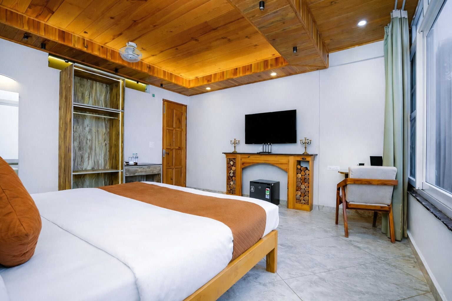 A bedroom with a bed, a TV mounted above a fireplace, a wooden wardrobe, and a chair by the window.