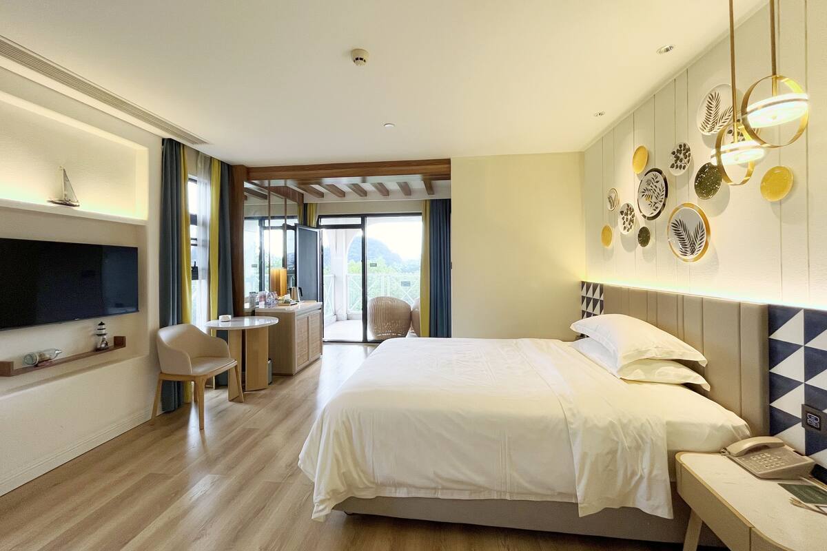 Quarto de hotel com cama, mesa de cabeceira, televisão e área de estar com poltrona e mesa.