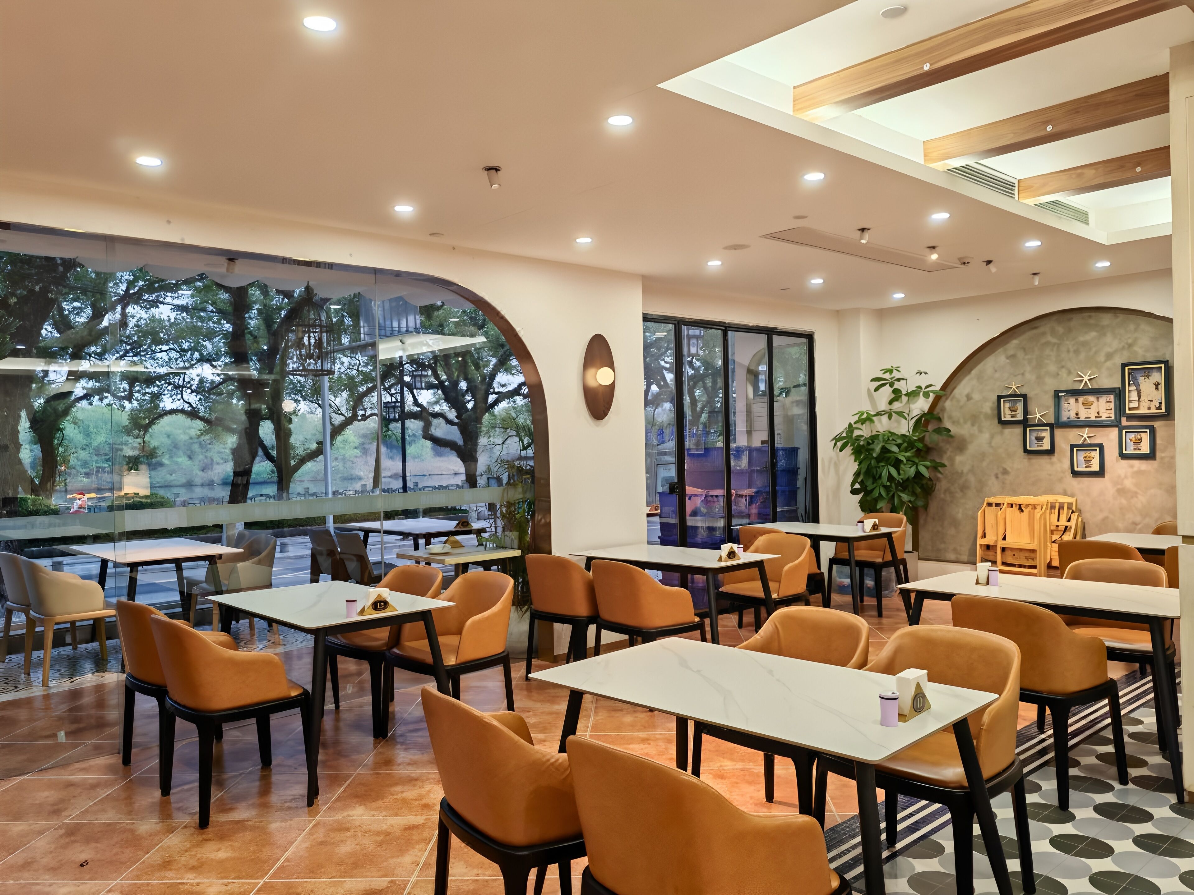 A modern dining area with tables and chairs, large windows with a view of trees, and a ceiling with wooden beams.