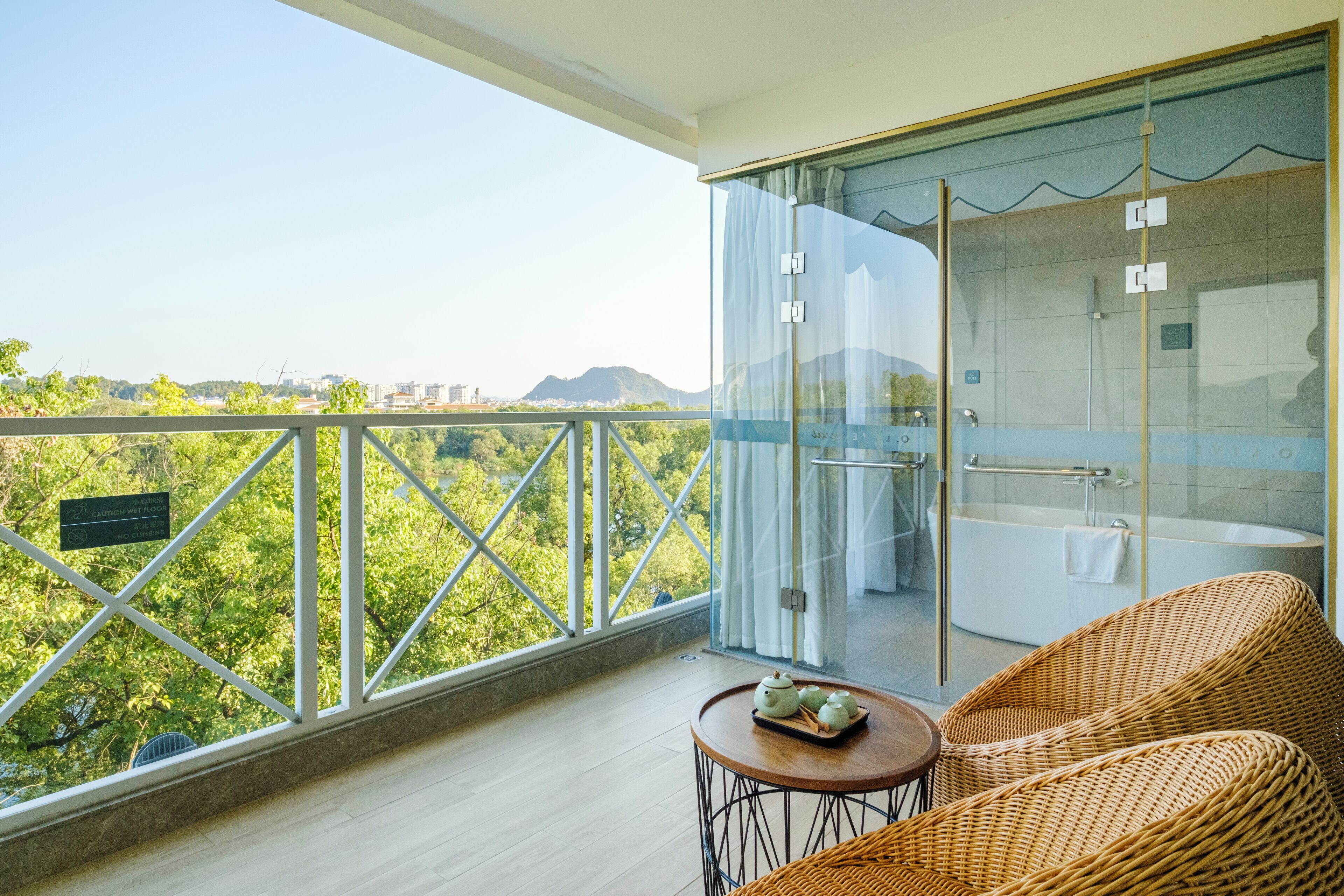 A balcony with a glass shower enclosure, wicker furniture, and a view of greenery and distant buildings.