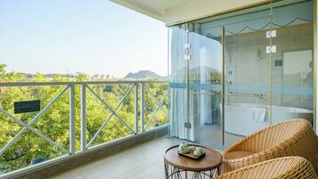A balcony with a glass shower enclosure, wicker furniture, and a view of greenery and distant buildings.