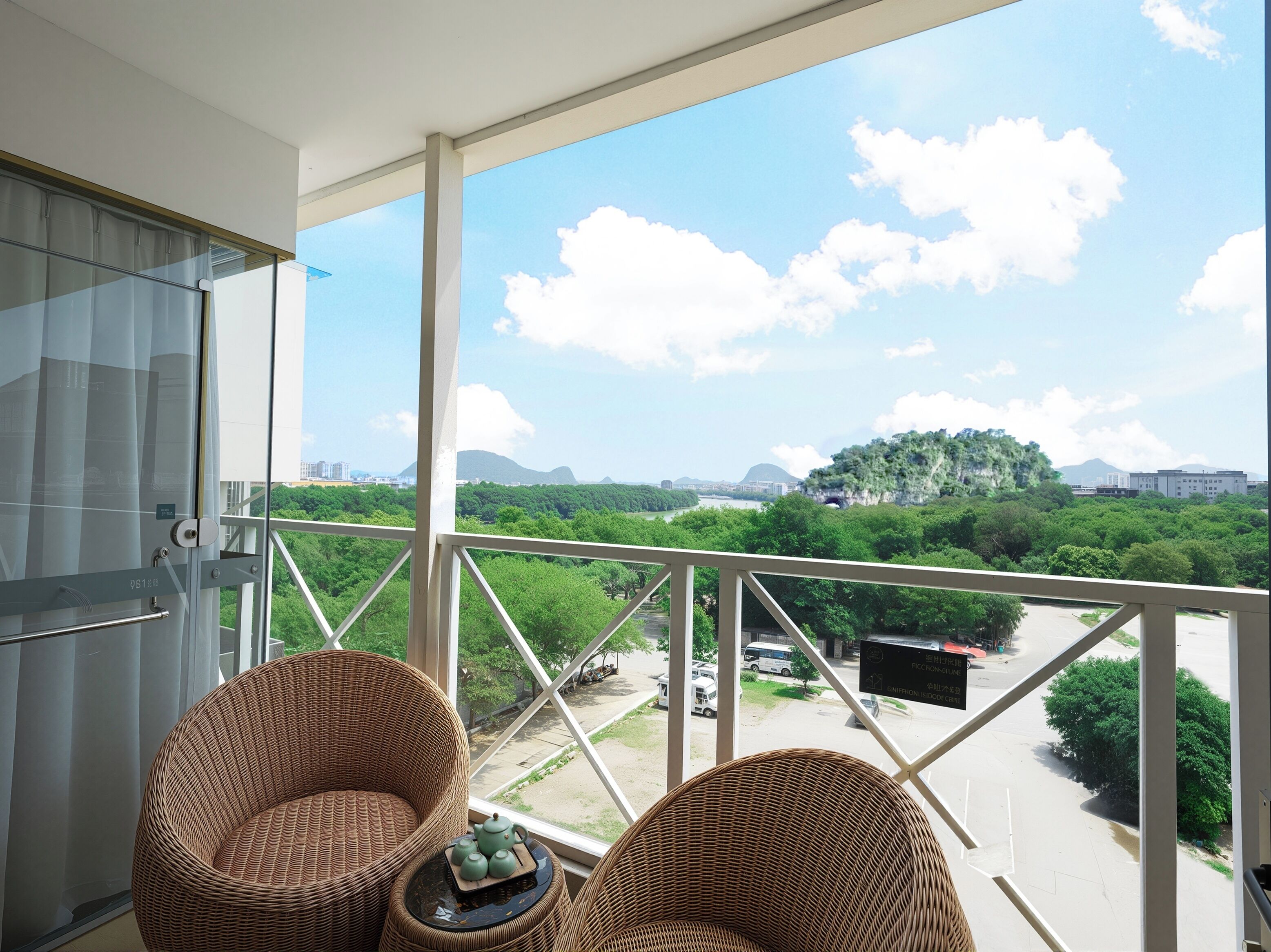 Balcón con sillas de mimbre y una mesa, con vistas a un paisaje urbano con áreas verdes y edificios.