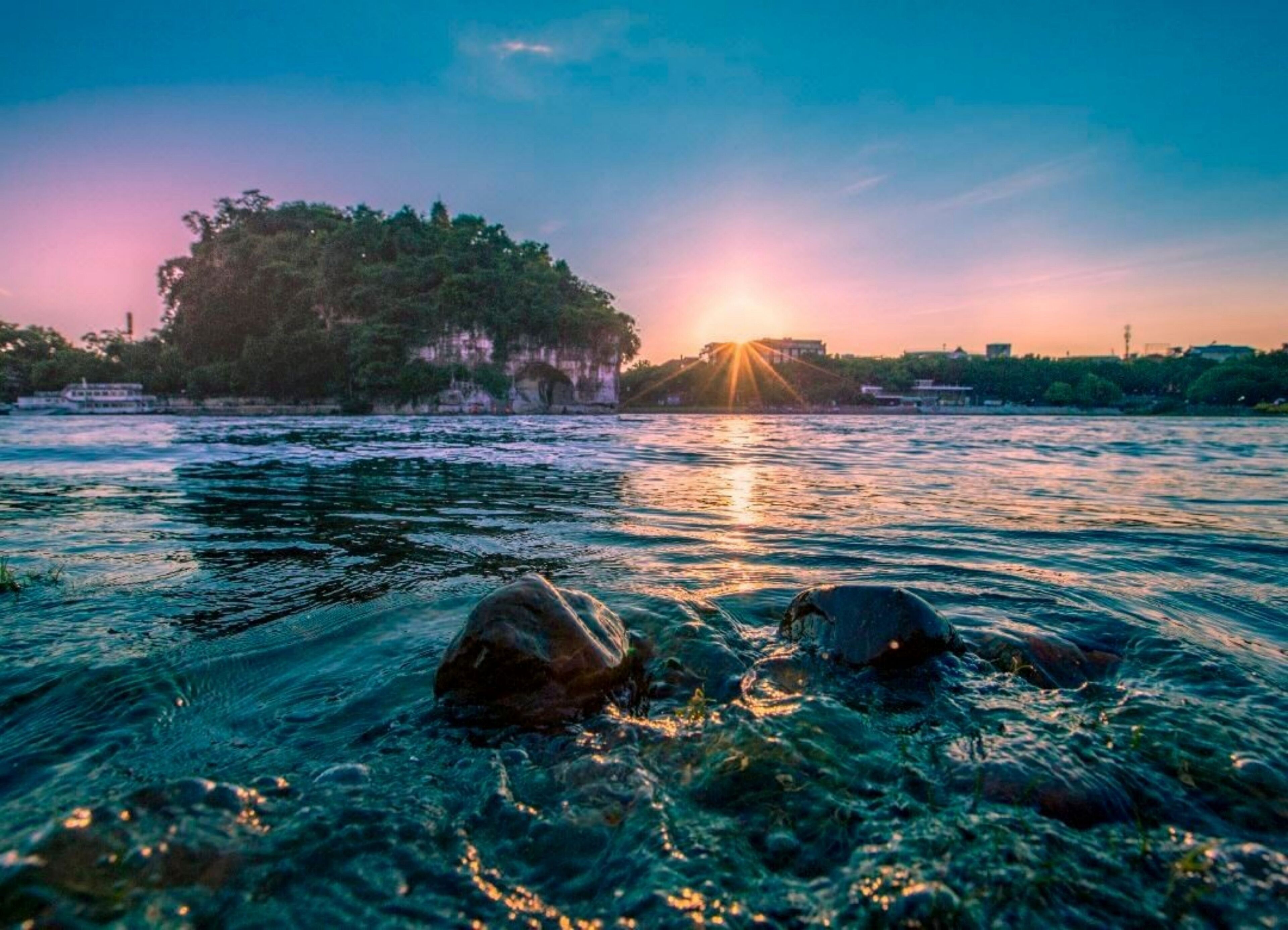 A river with clear water, rocks in the foreground, and a sunset in the background.