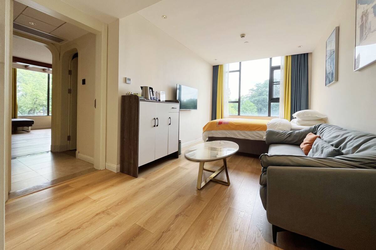 Quarto de hotel moderno com sofá, uma pequena mesa redonda, cama com cobertor amarelo, televisão e guarda-roupa.