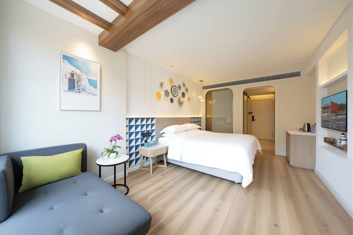 Quarto de hotel moderno com uma cama, um sofá, uma mesa pequena com uma planta e uma TV na parede.