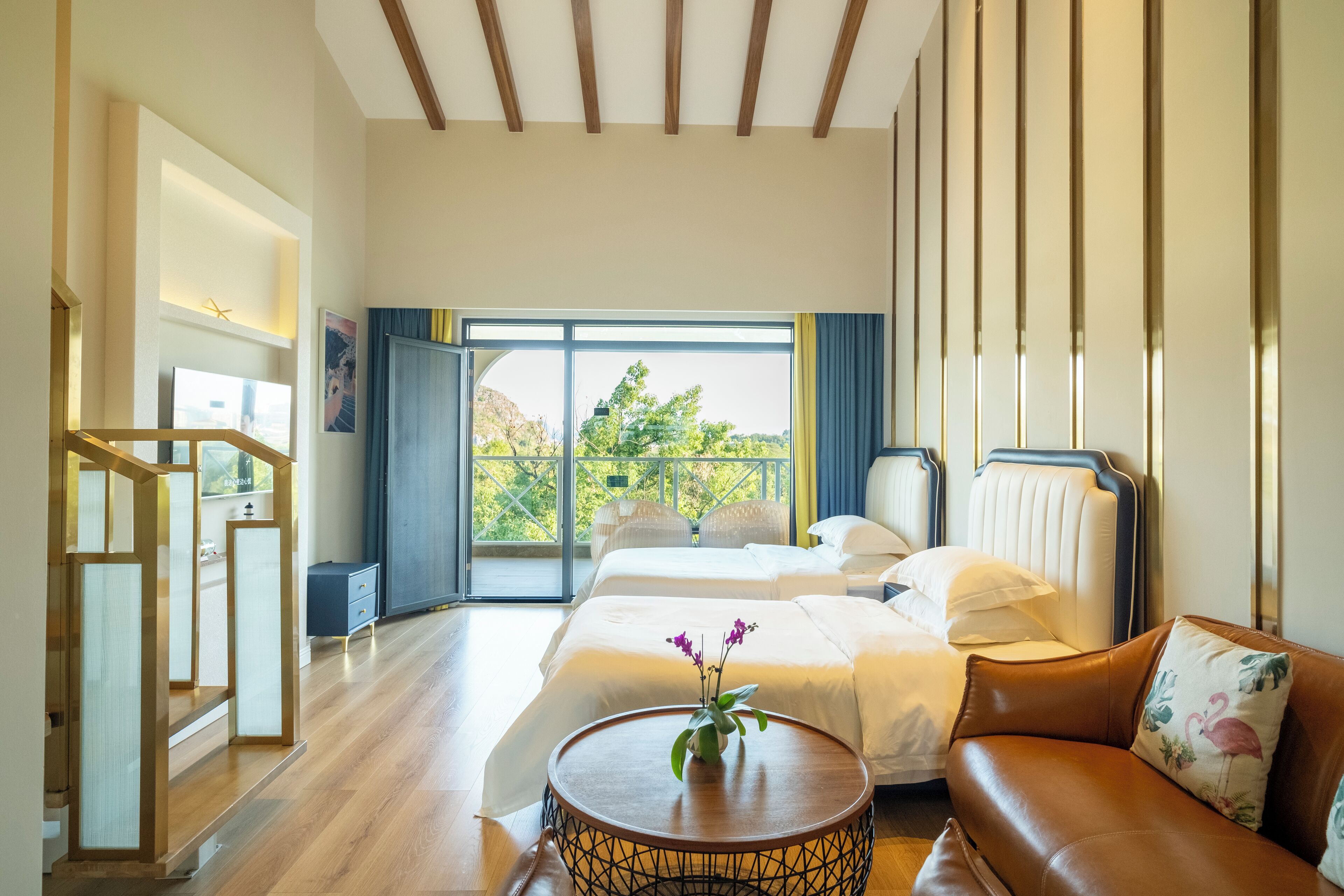 A hotel room with two beds, a sofa, a round coffee table with flowers, a balcony with a view of greenery, and a wooden staircase.
