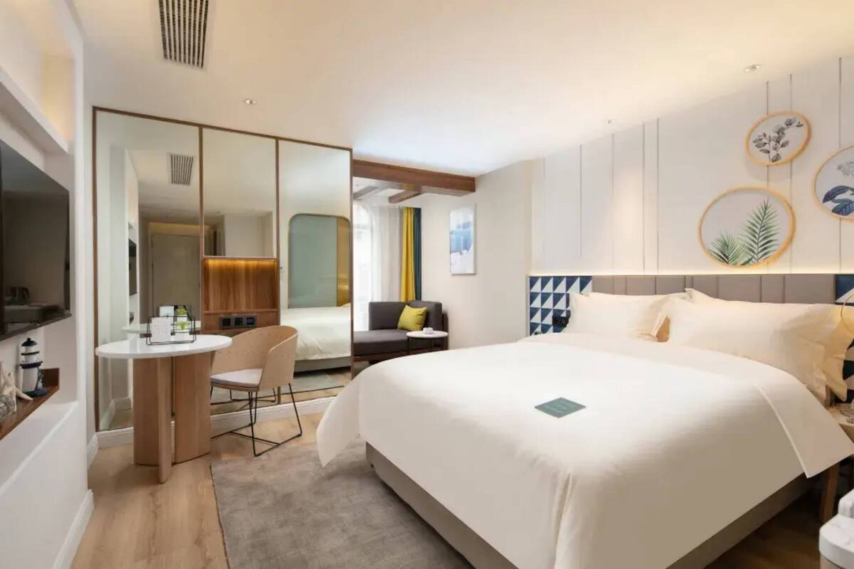 Quarto de hotel moderno com uma cama grande, uma escrivaninha com cadeira, uma mesinha e um espelho.