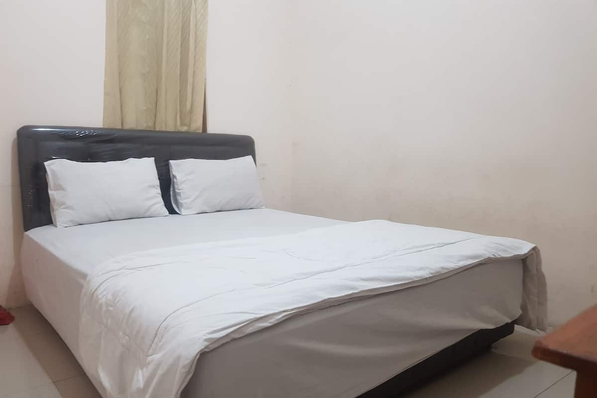 Uma cama de solteiro com roupa de cama branca e cabeceira escura num quarto com paredes lisas e chão em azulejos.