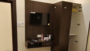 A modern hotel room with a large bed, a flat-screen TV mounted on the wall, a wardrobe, and a small seating area. - Moonvilla Banquet And Hotels (Phulpur)