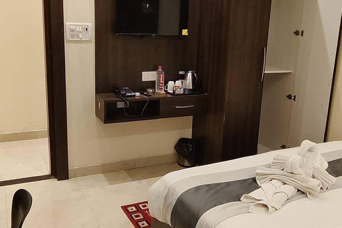A modern hotel room with a large bed, a flat-screen TV mounted on the wall, a wardrobe, and a small seating area.