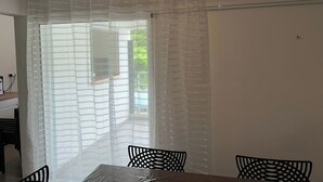 Ein Zimmer mit Esstisch und Stühlen, einem Fenster mit durchsichtigen Vorhängen und Blick ins Grüne.