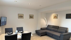 Een moderne woonkamer met een eethoek, een grijze bank en een flatscreen televisie aan de muur.