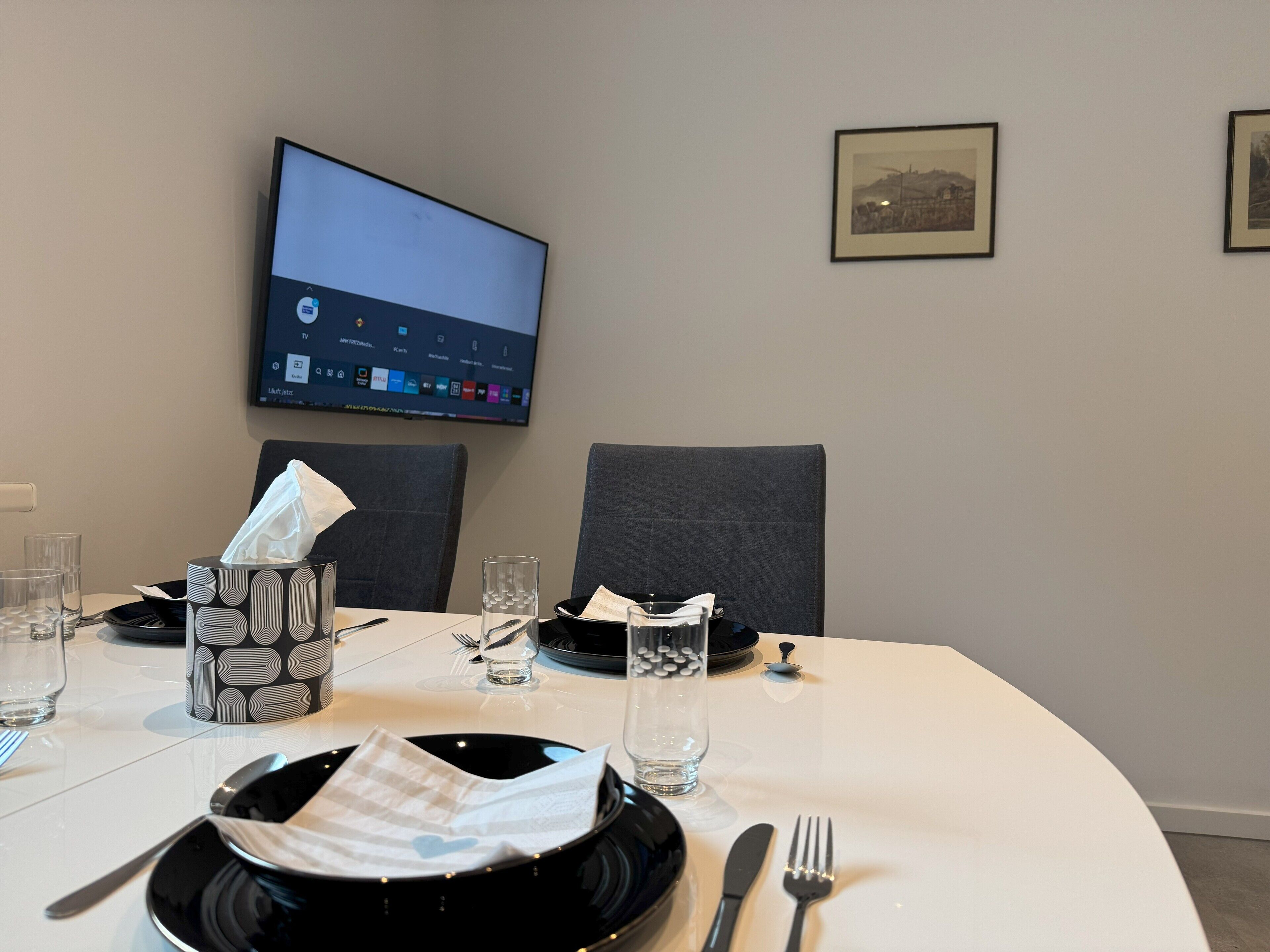 A hotel room with a neatly set dining table, a flat-screen TV mounted on the wall, and framed pictures on the wall.