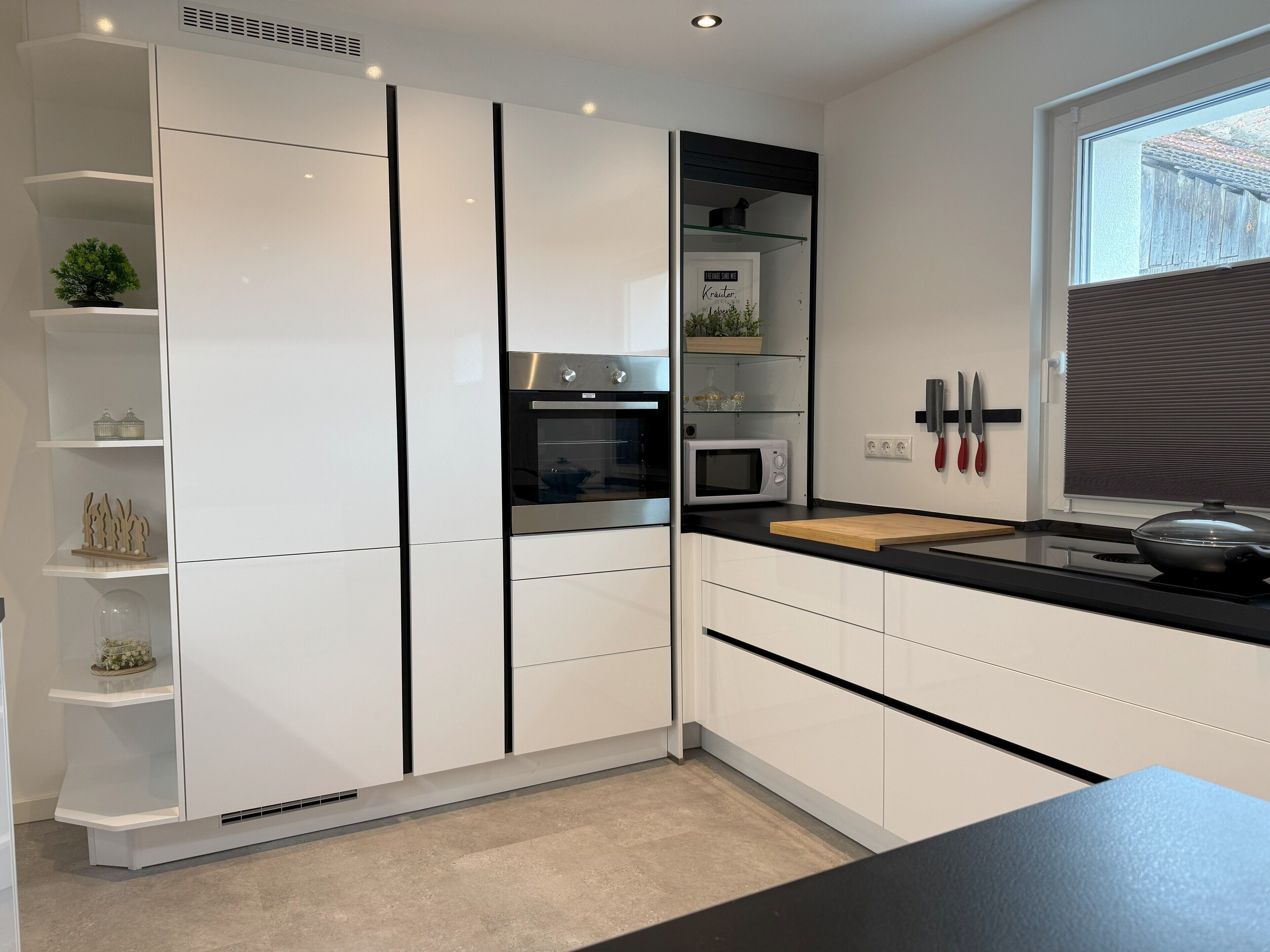 A modern kitchen with white cabinetry, a black countertop, a built-in oven, a microwave, and a window with blinds.