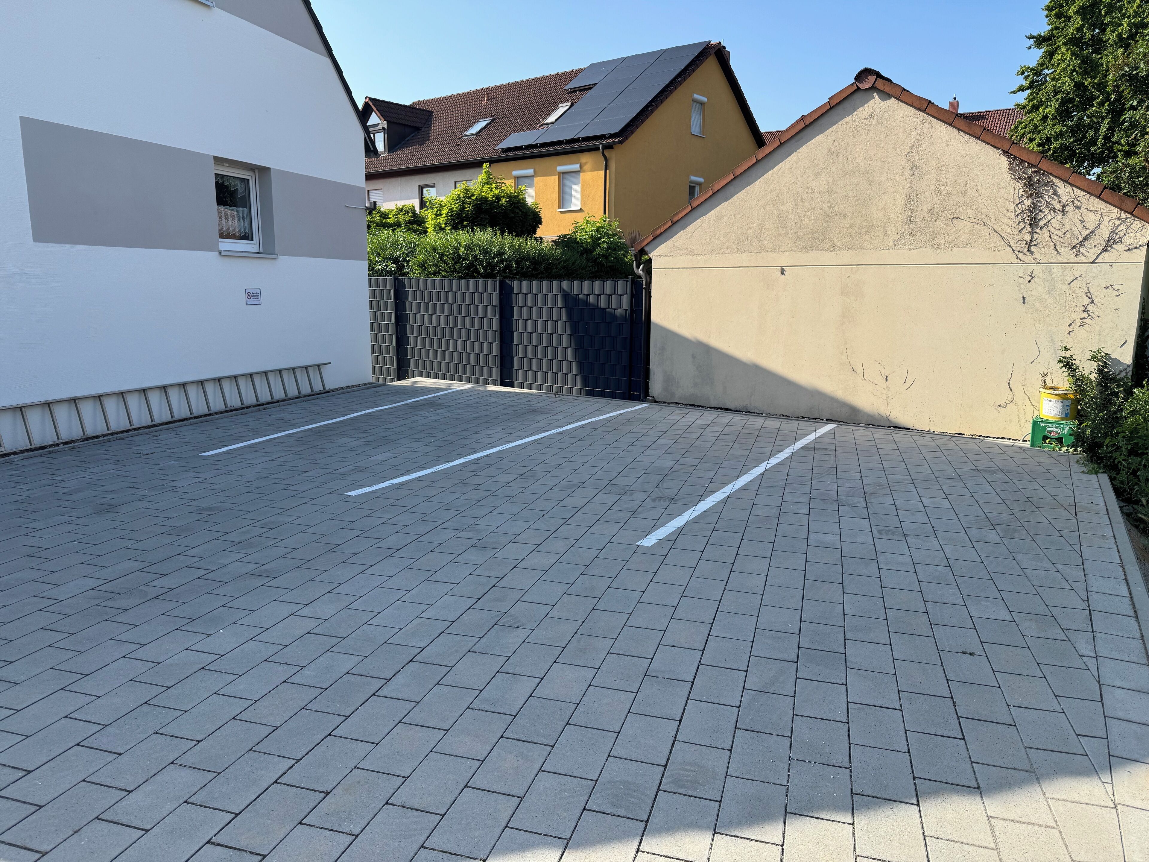 Ein gepflasterter Parkplatz mit markierten Stellplätzen, einem befahrten Durchgang und Wohnhäusern im Hintergrund.