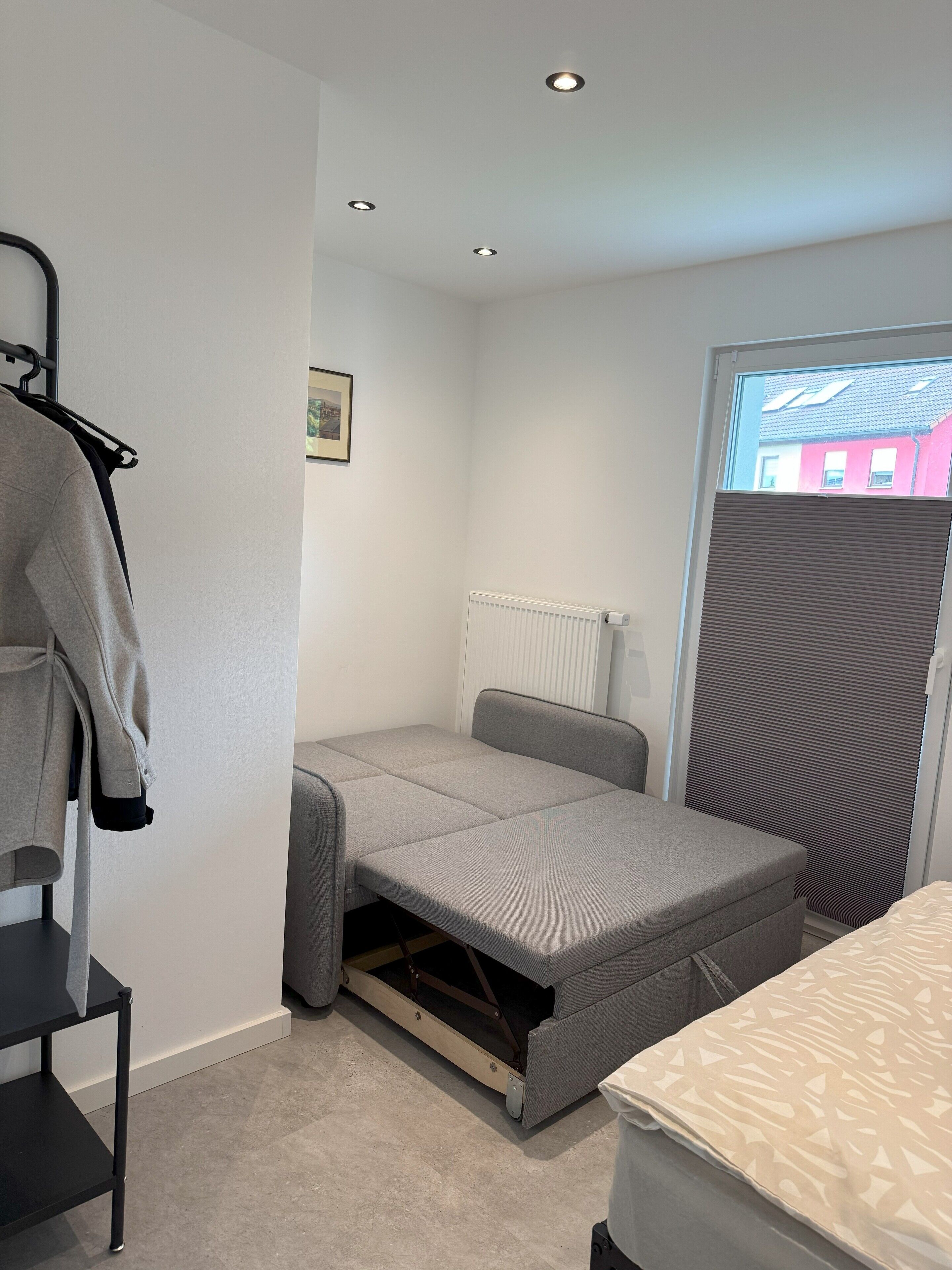 A compact room with a pull-out bed, a radiator, a window with blinds, and a coat rack with a jacket.
