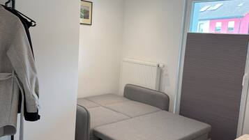 A compact room with a pull-out bed, a radiator, a window with blinds, and a coat rack with a jacket.