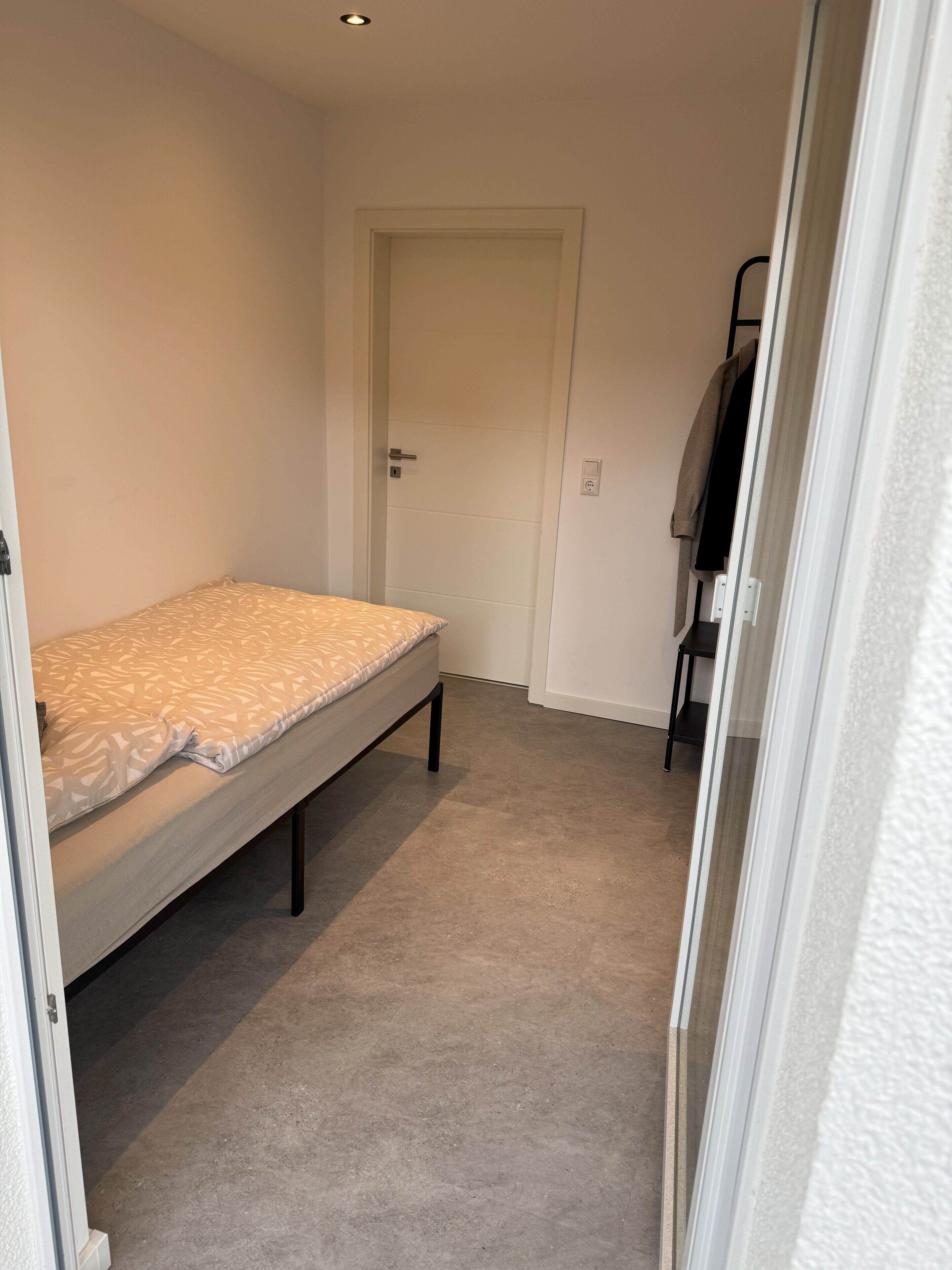A small, narrow room with a single bed, a door, and a small table with a chair.