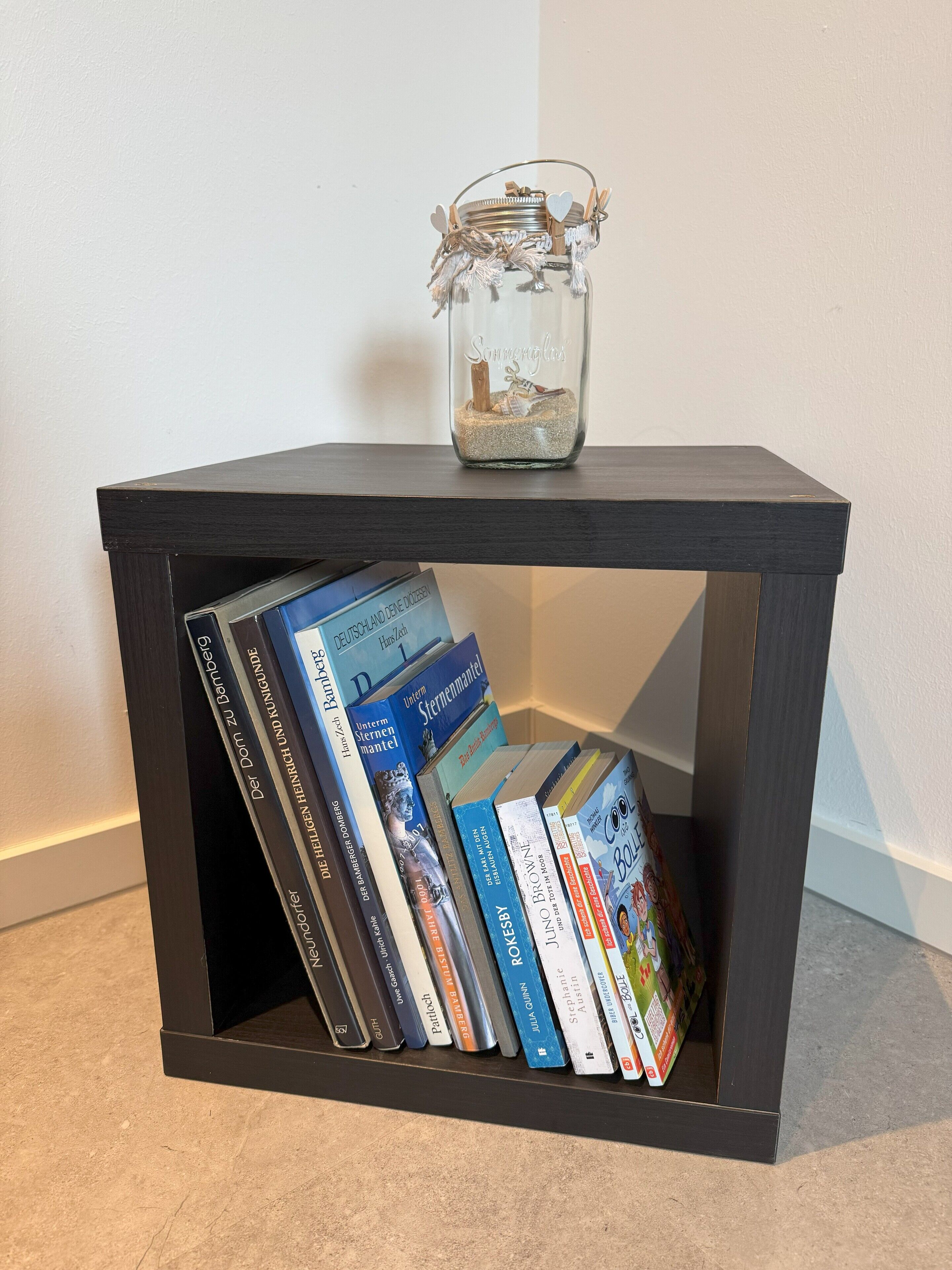 A small black side table with a glass jar containing sand and a stick, and a row of books underneath.