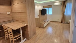A modern living room with a wooden floor, a built-in TV, a dining area with chairs, and a window with curtains. - Simmi 4 Hotel & Apartment Near SECC (Ho Chi Minh City)