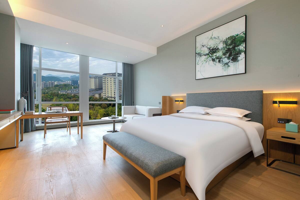 Habitación de hotel moderna con una cama grande, un escritorio, un banco y vistas de la ciudad a través de amplios ventanales.