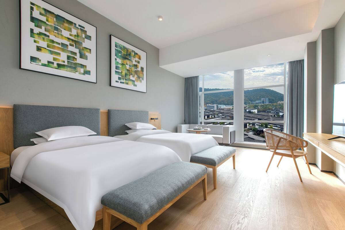 Habitación de hotel con dos camas, un escritorio, una silla y grandes ventanas con vistas a la ciudad.
