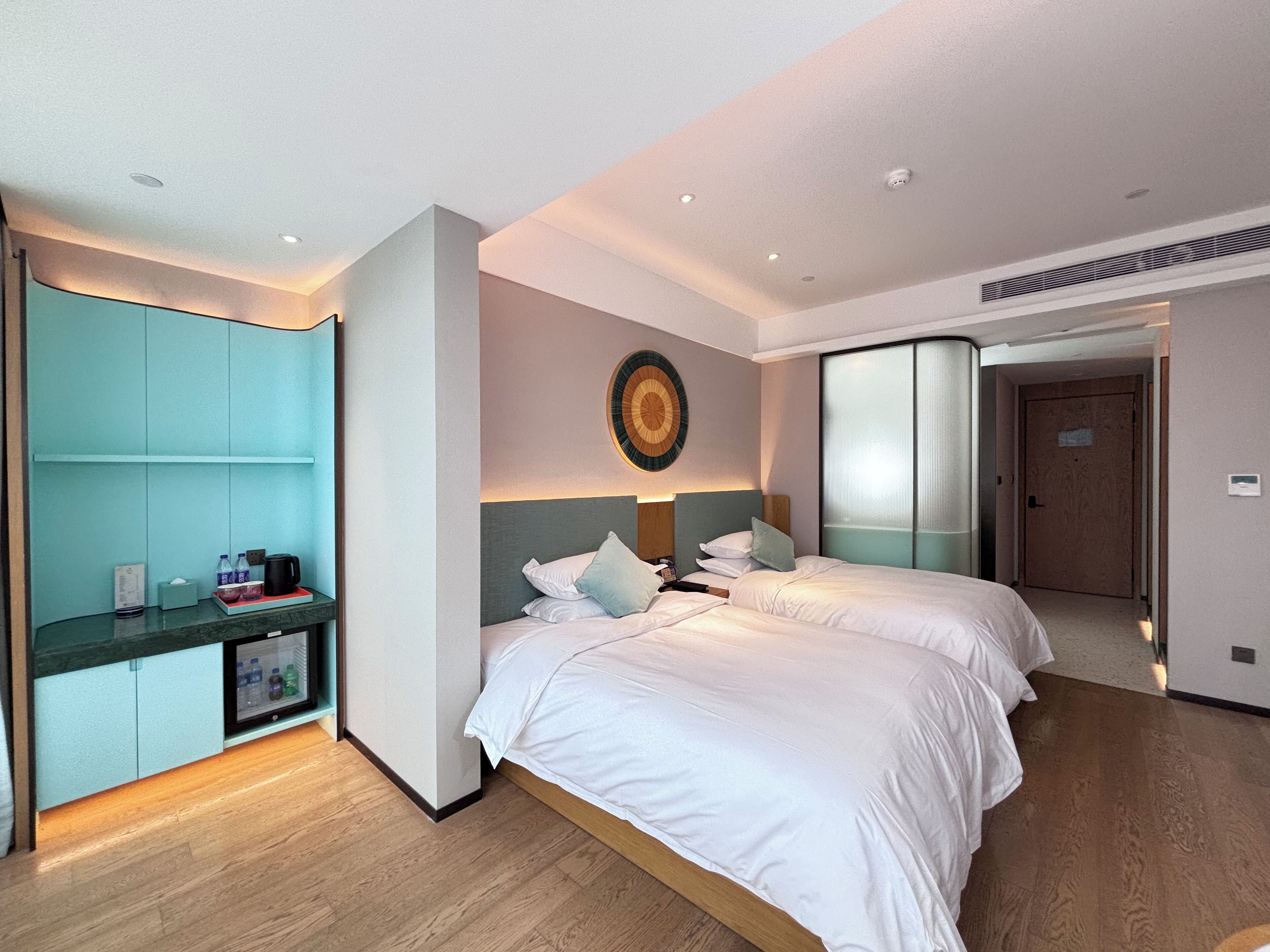 Une chambre d’hôtel avec deux lits, un minibar, une horloge murale et une porte coulissante donnant sur une autre chambre.