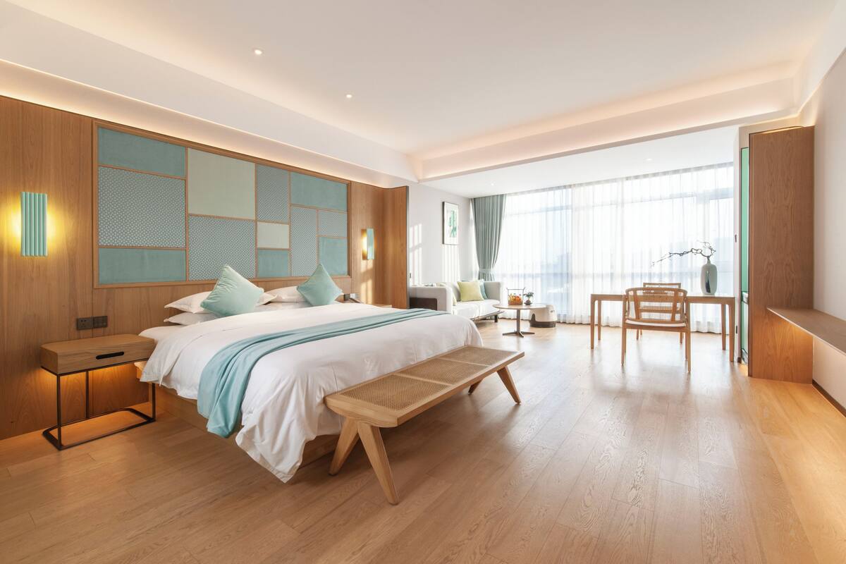 Habitación de hotel moderna con una cama grande, un banco de madera, un escritorio y vistas de la ciudad a través de amplias ventanas.