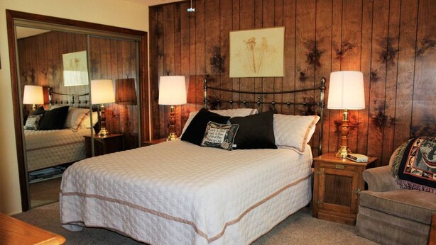 A bedroom with a large bed, two bedside tables with lamps, a wooden headboard, and a mirror. - Cabin with Foosball-Southern Comfort (Big Bear Lake)