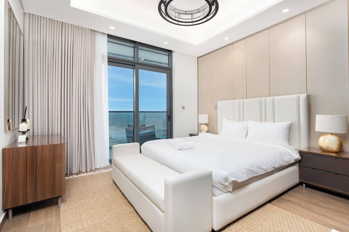 Un dormitorio moderno con una cama grande, una mesita de noche, un aparador de madera y vista al mar a través de un ventanal.