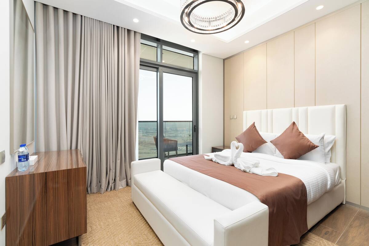 Habitación de hotel moderna con una cama grande, una mesita de noche de madera, un banco y vistas al mar a través de un ventanal amplio.