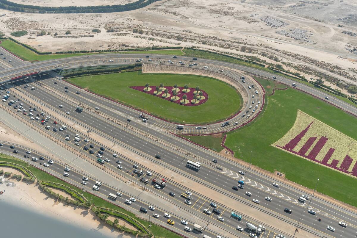 Vista aérea de un intercambio de autopistas con múltiples carriles y una zona ajardinada con árboles y flores.