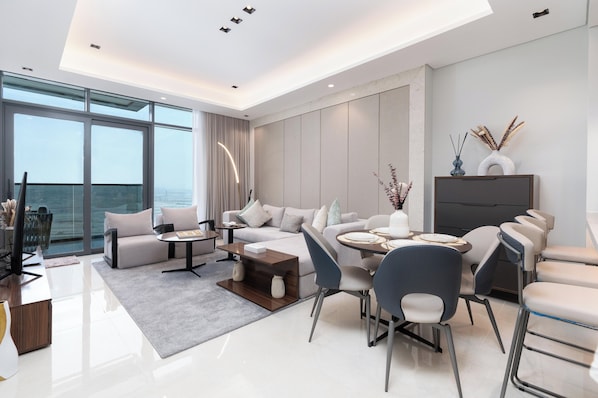 A modern living room with a sofa, coffee table, and chairs, a television, and a large window with a view of the sea. - Samaya Holiday Homes Dubai Luxury Suites (Dubai)