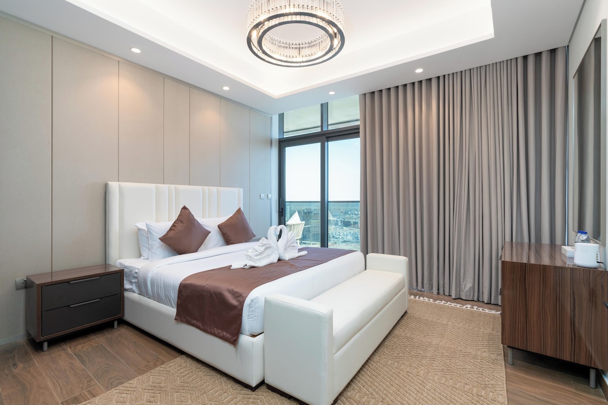 Ein modernes Hotelzimmer mit einem großen Bett, einem Holfnachtkästchen, einem Schreibtisch und Blick auf die Stadt durch große Fenster.