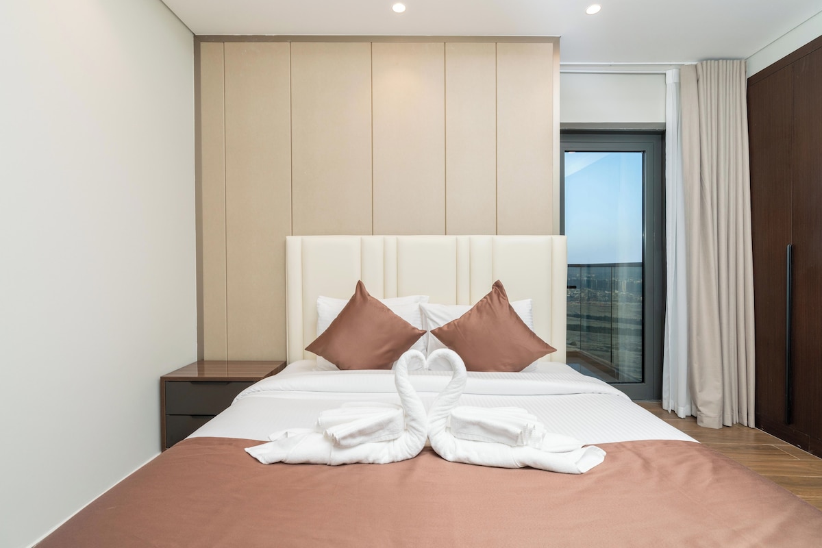 Ein Hotelzimmer mit einem großen Bett, zwei Kissen, einem Nachttisch und Blick auf die Stadt durch eine Glastür.