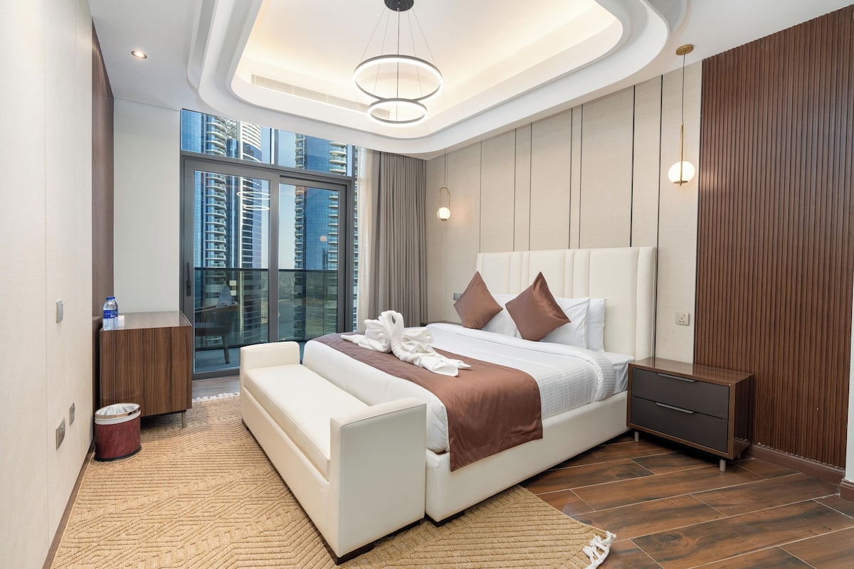 Ein modernes Schlafzimmer mit einem großen Bett, einem Nachttisch, einer Ablage, einer Couch, einem Mülleimer und Blick auf die Stadt durch große Fenster.