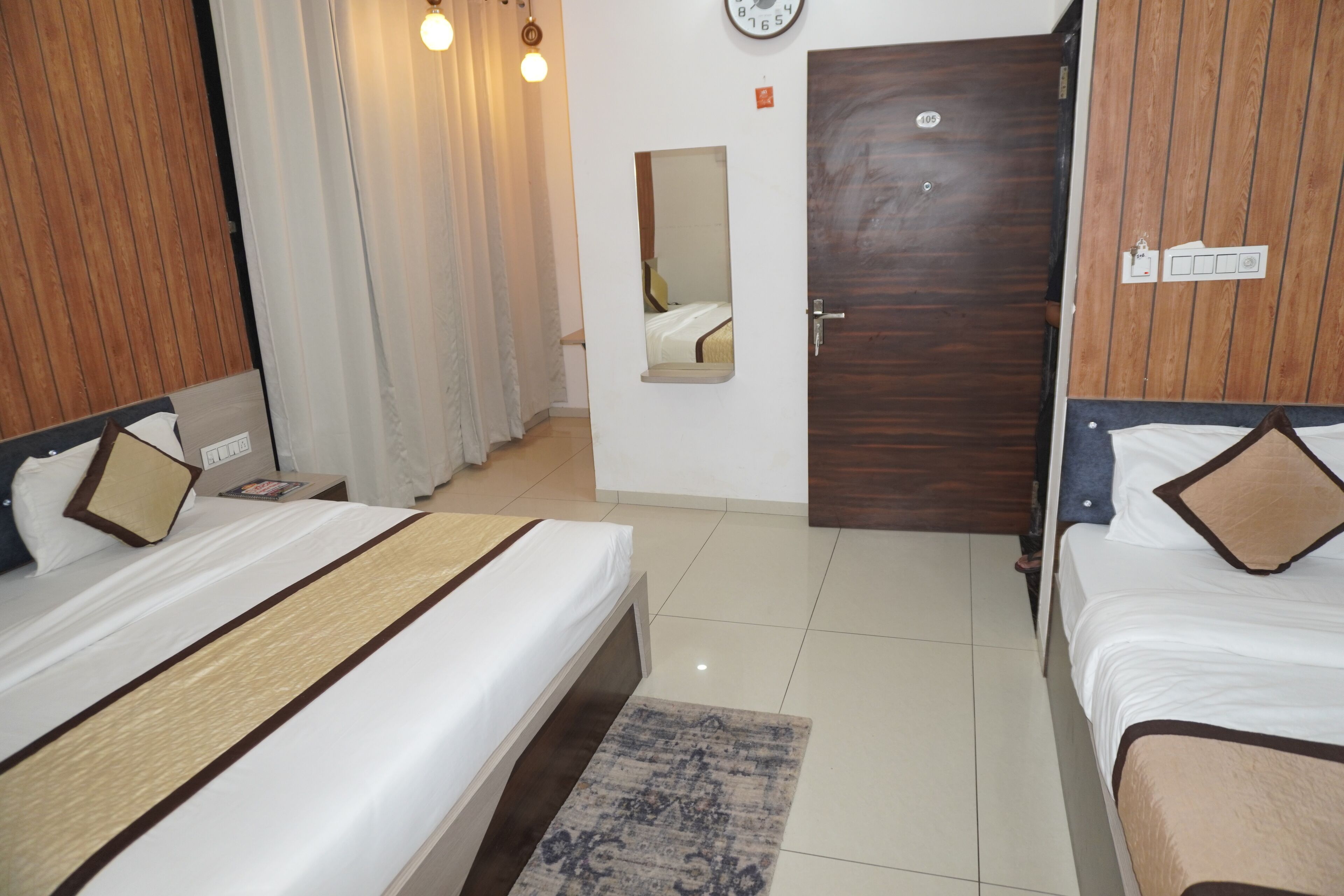 A hotel room with two beds, a wooden wardrobe, a mirror, a clock, and a door with a number plate.