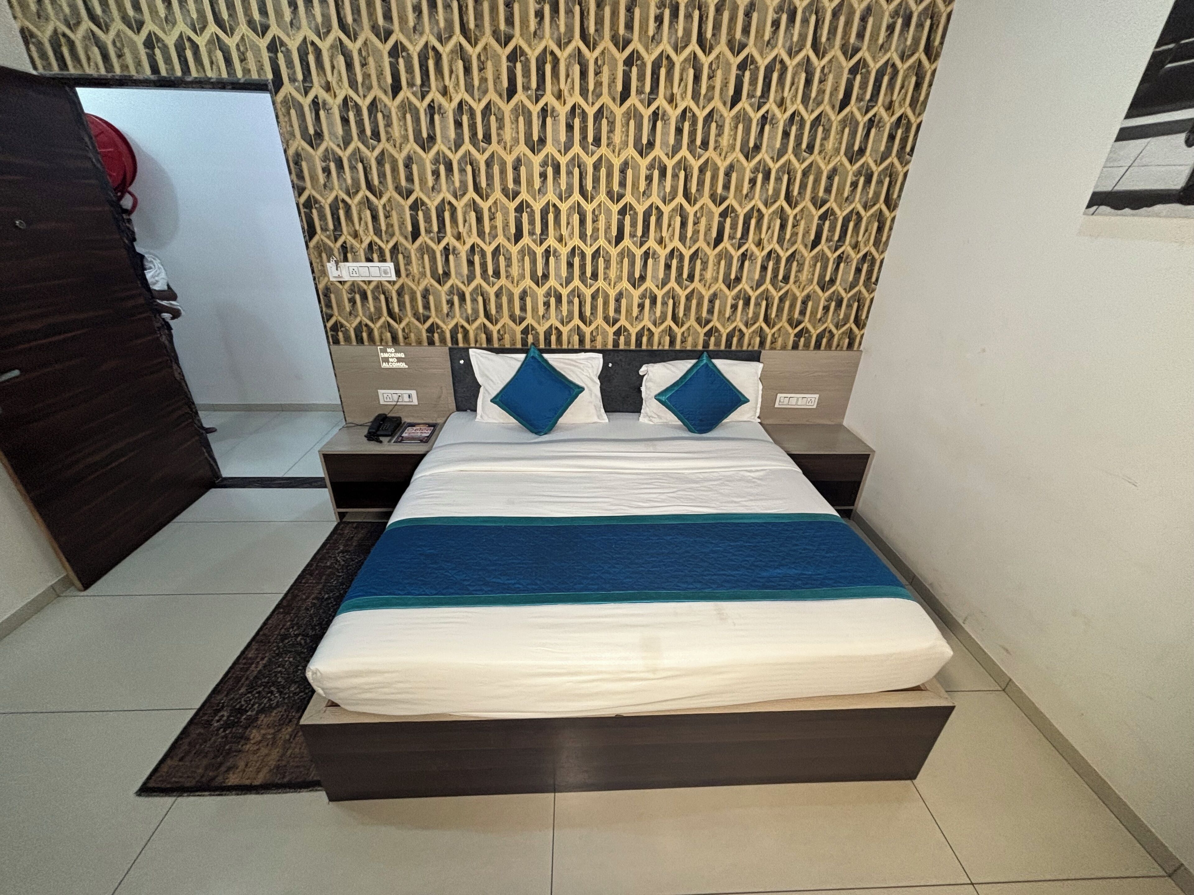 Un lit bien fait, recouvert d’une couvre-lit bleue et blanche, deux tables de chevet avec des lampes, un mur à motifs et une porte sur le côté gauche.