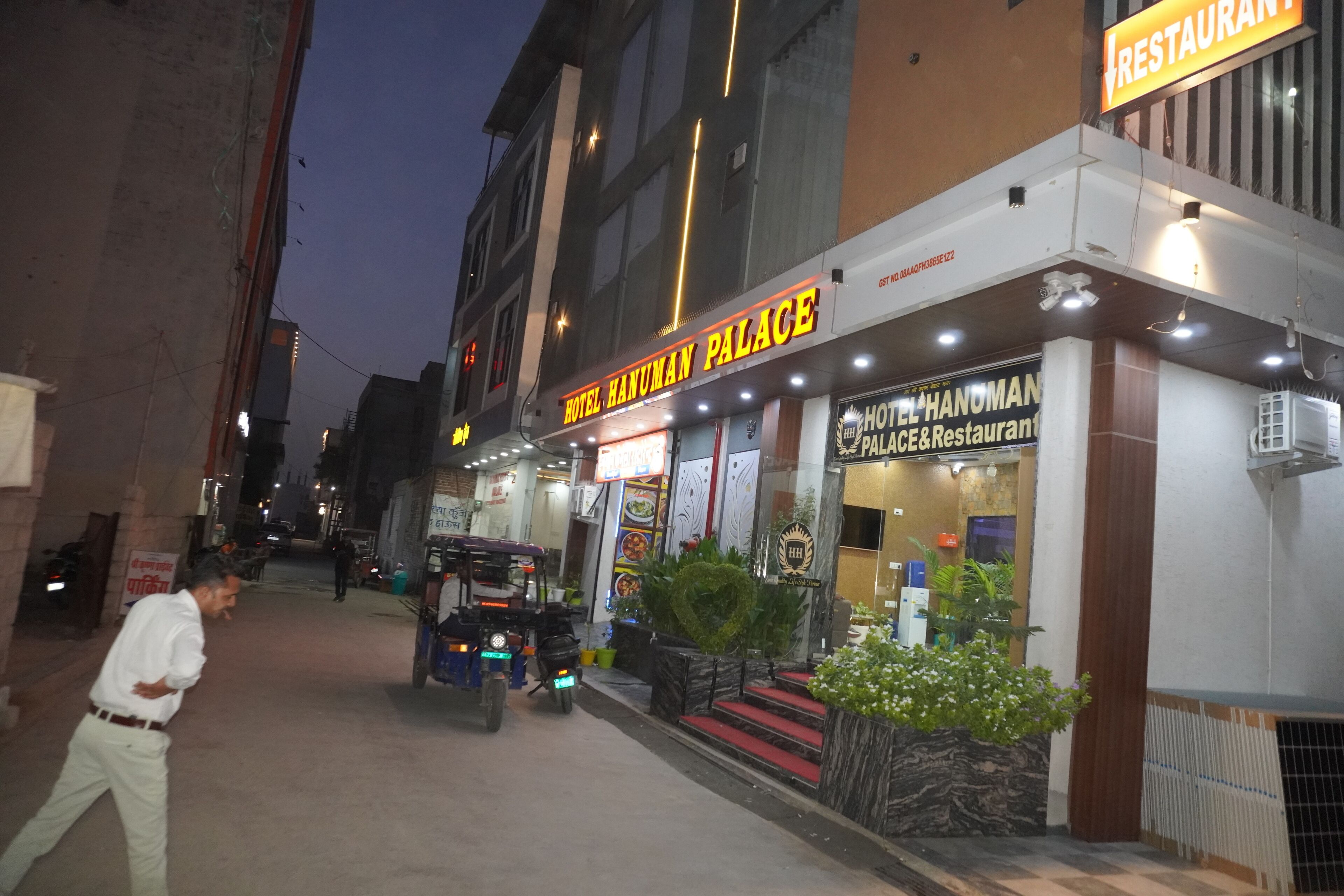 A hotel named Hotel Hanuman Palace with a restaurant sign, a parked three-wheeler, and a person walking by.