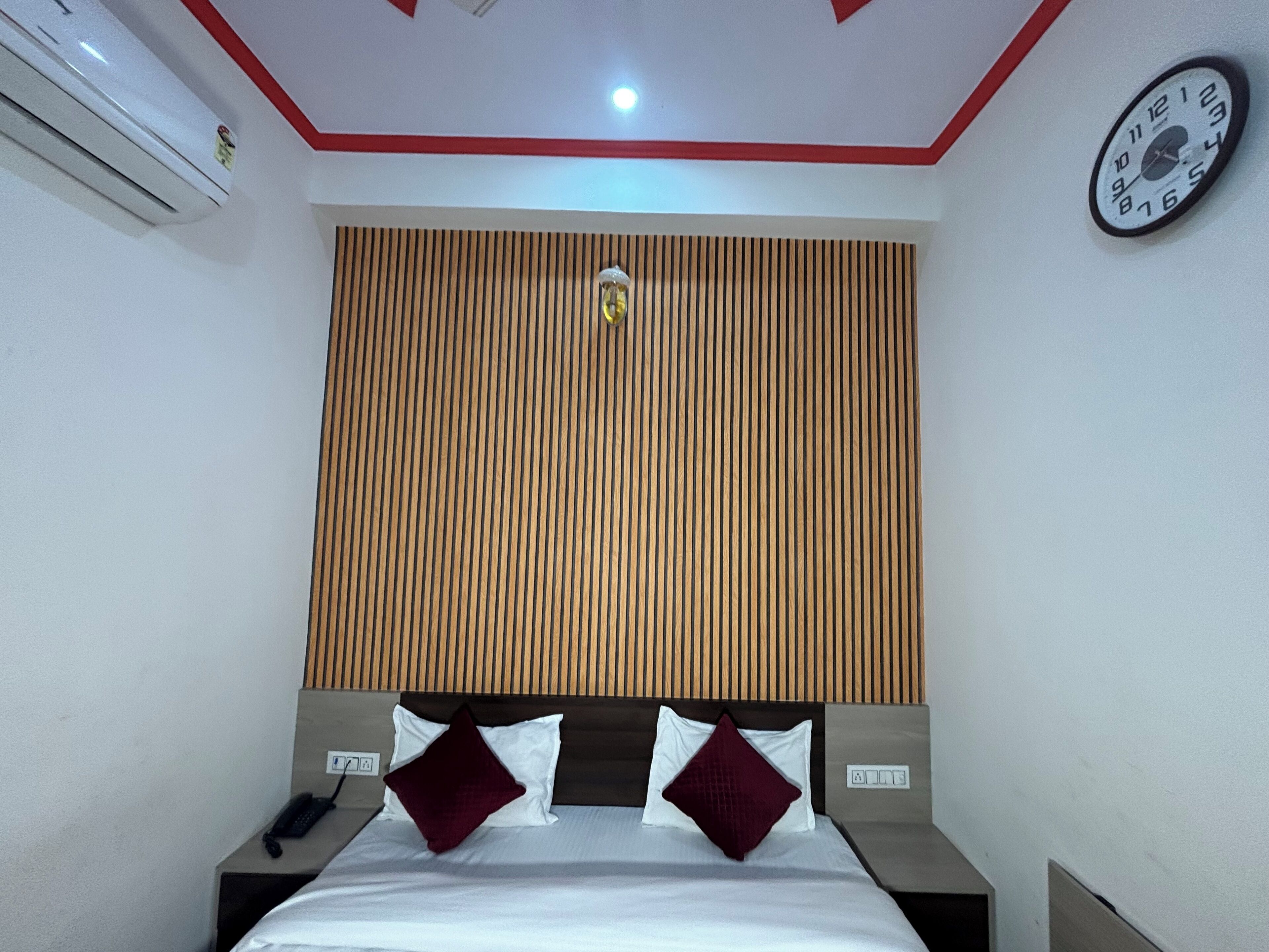 A hotel room with a bed, a wall clock, a telephone, and a wall-mounted air conditioner.