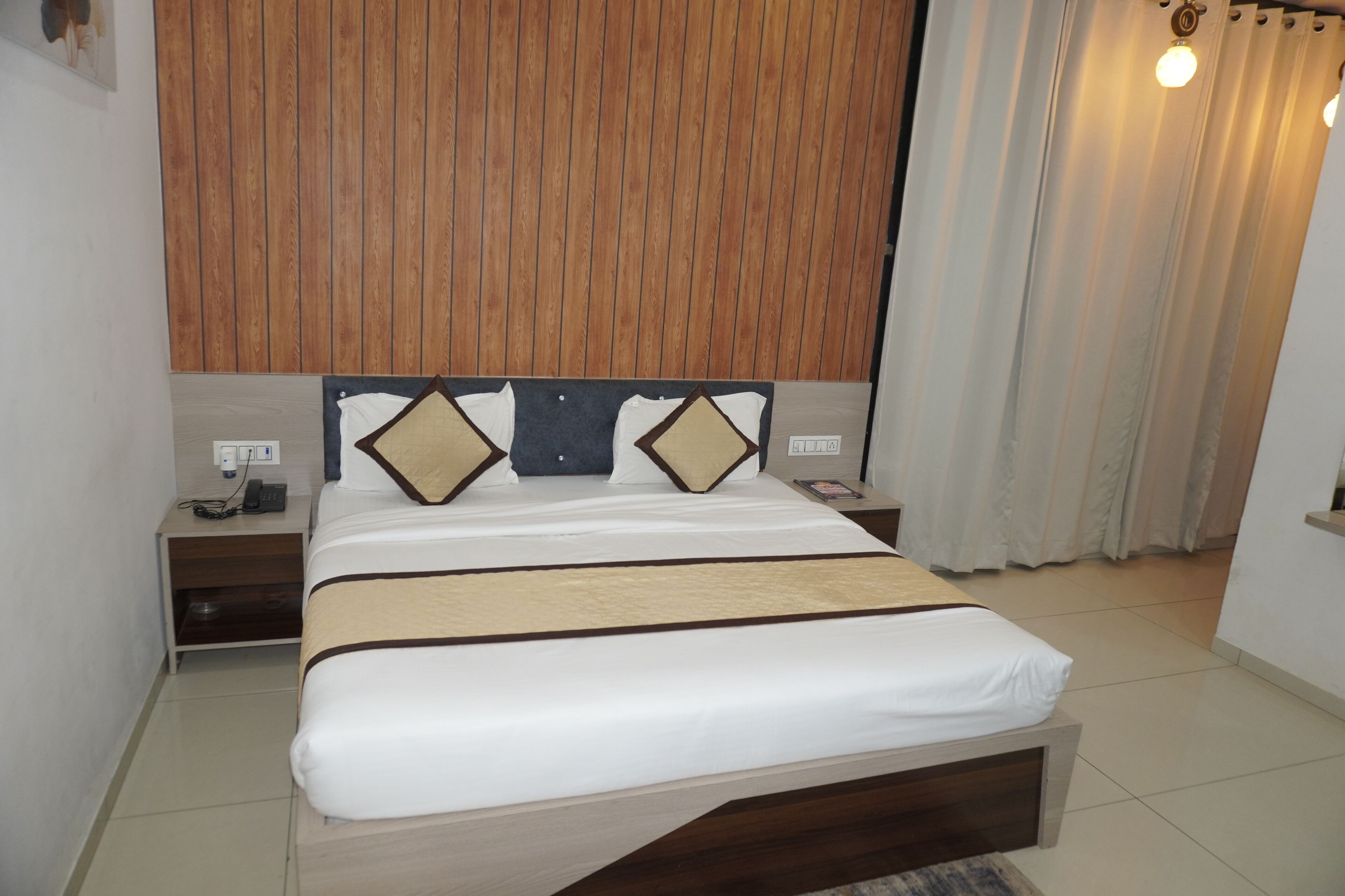 A neatly made bed with a wooden headboard, a bedside table with a lamp, and a curtain to the right.