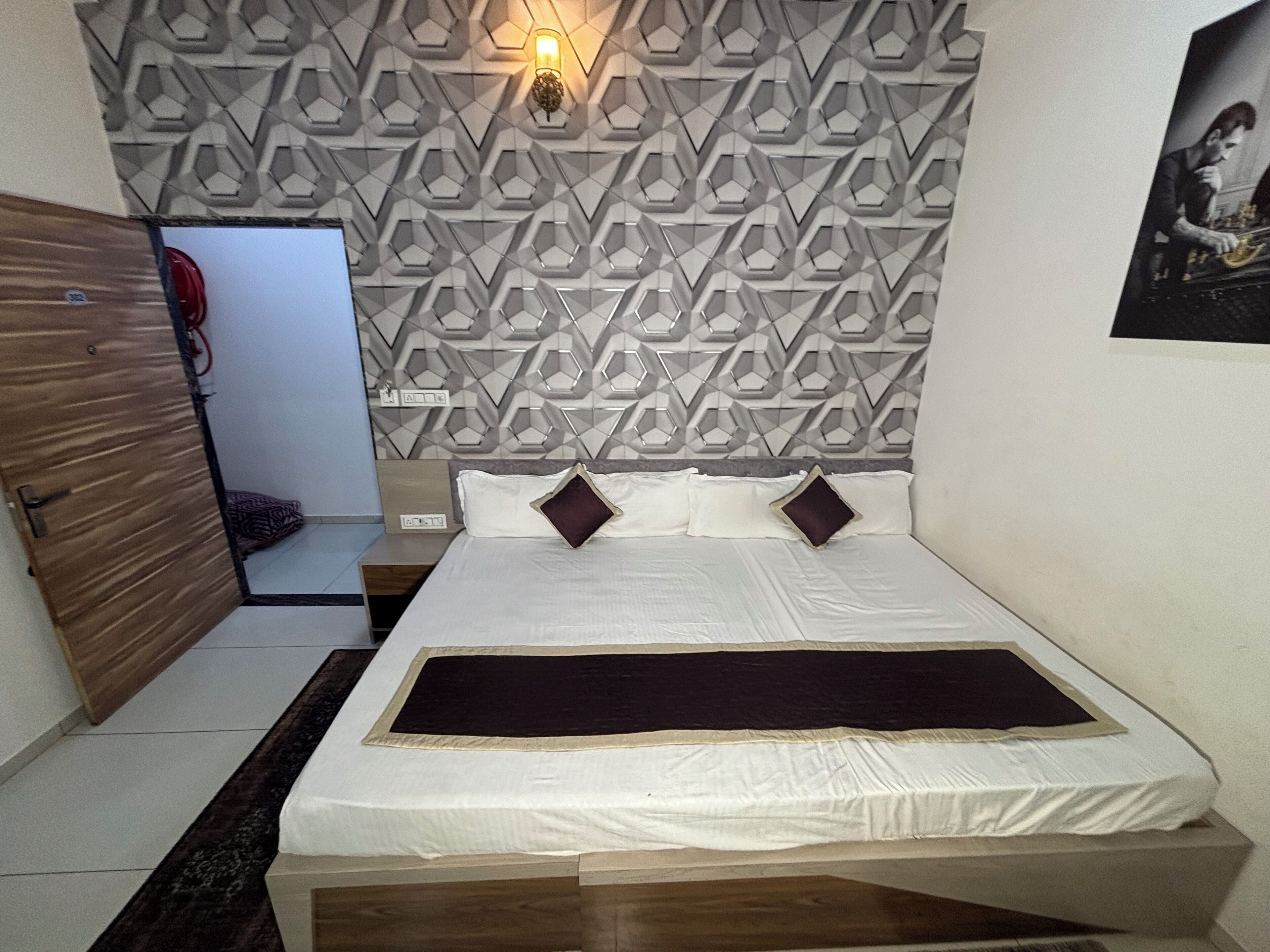 A bedroom with a geometric patterned headboard, a bed with white linens and two dark pillows, a wooden door, and a small bedside table.