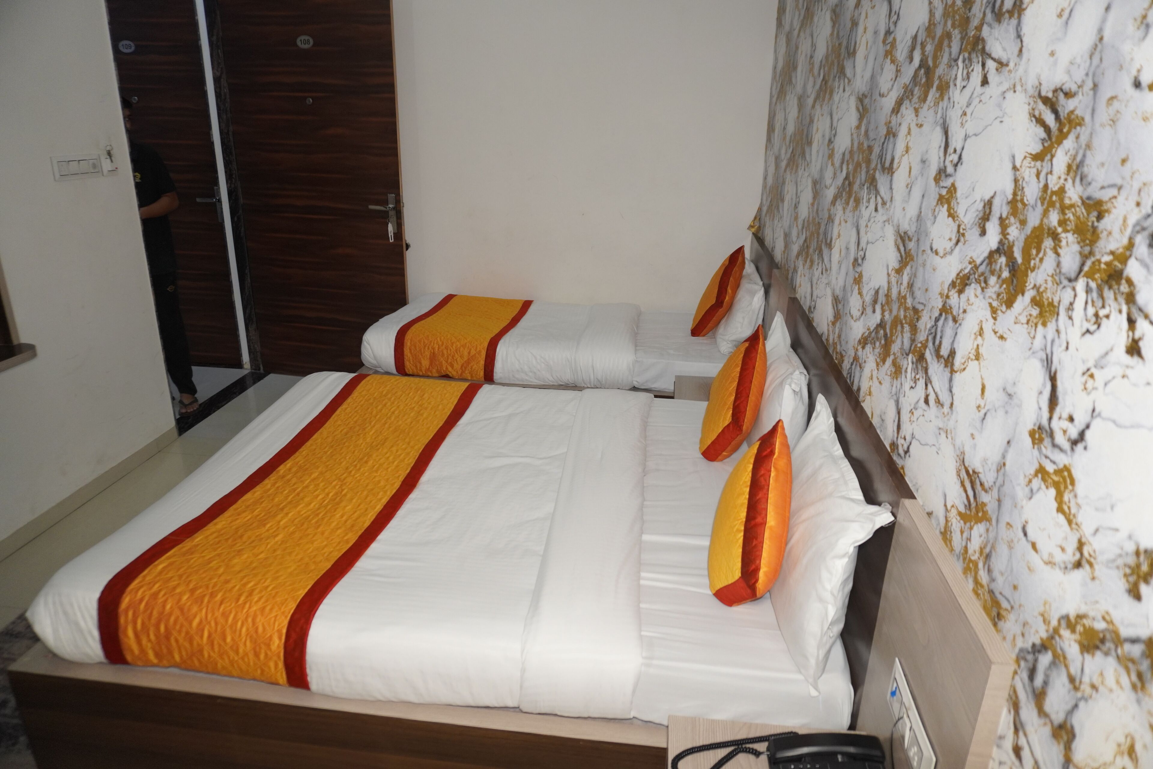 A hotel room with two beds, each with white bedding and orange accents. There is a wooden headboard and a marble-patterned wall.