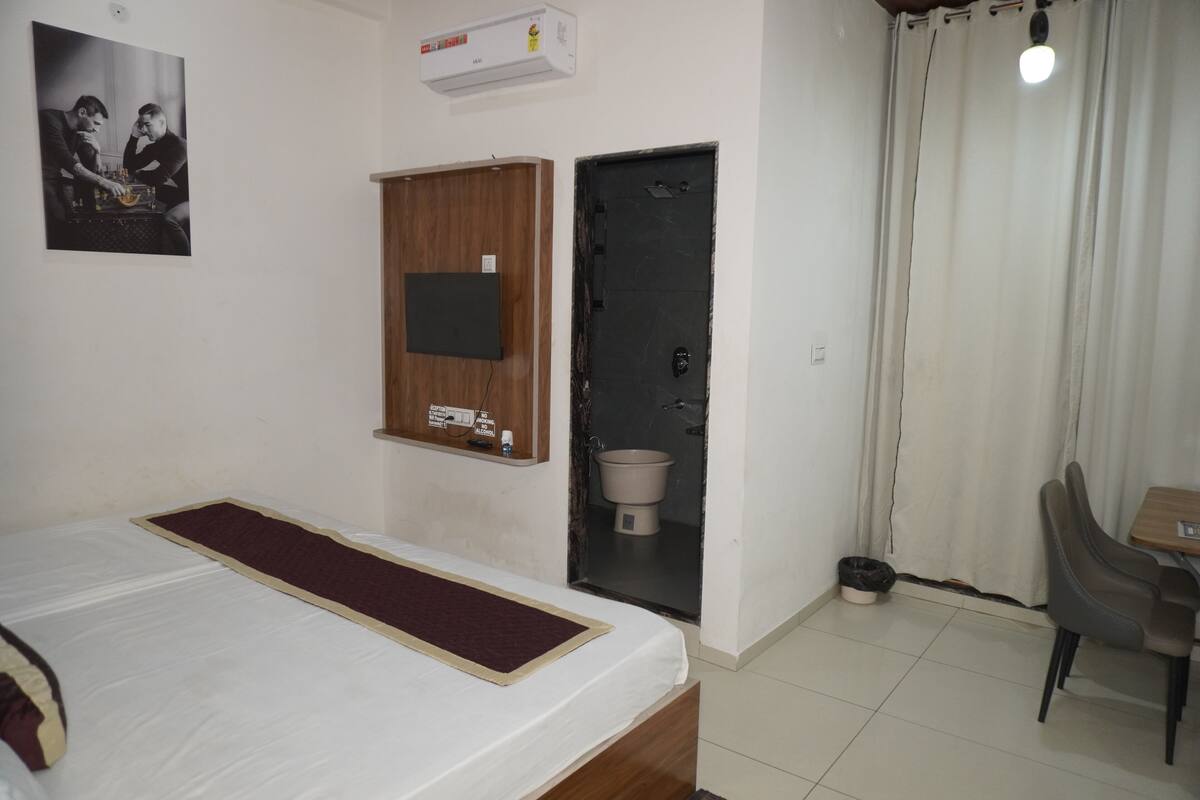 Habitación de hotel con cama, televisor montado, mueble de madera, puerta, mesita pequeña y una silla.